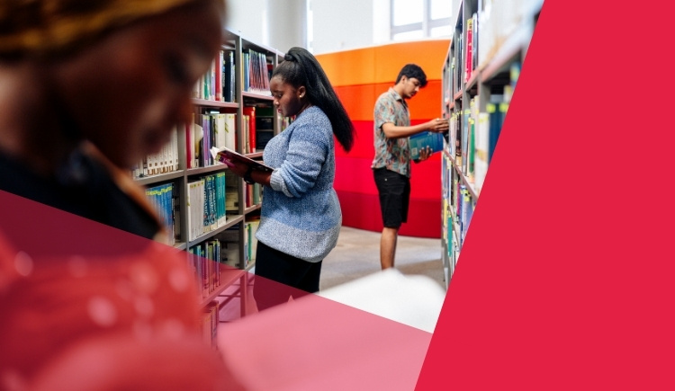 People browsing bookshelves in a library, with one person in the foreground looking down at a book and two others in the background selecting books.
