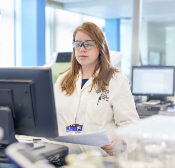 A female student wearing white lab coat and goggles looking at computer screen in lab.