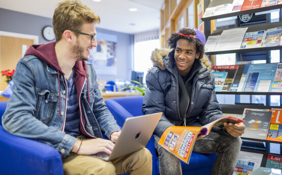 Two men talking, one using a laptop, one reading a magazine