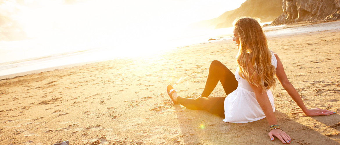 Person sitting on a bench, by a tree on a beach. 