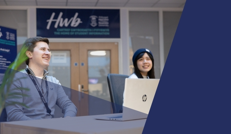 Two staff members sit behind a desk with a laptop in front of them. A sign that reads 