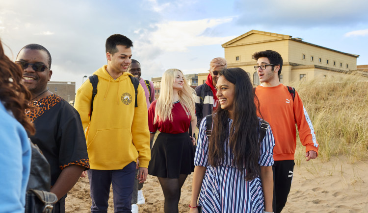 The image shows a group of students walking to the beach. Behind them is The Great Hall on Bay Campus.