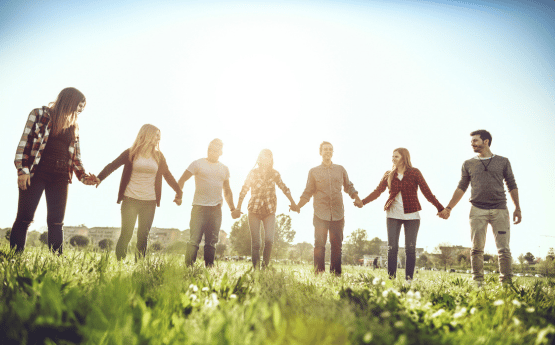A group of people holding hands in a field with the sunshine behind them.
